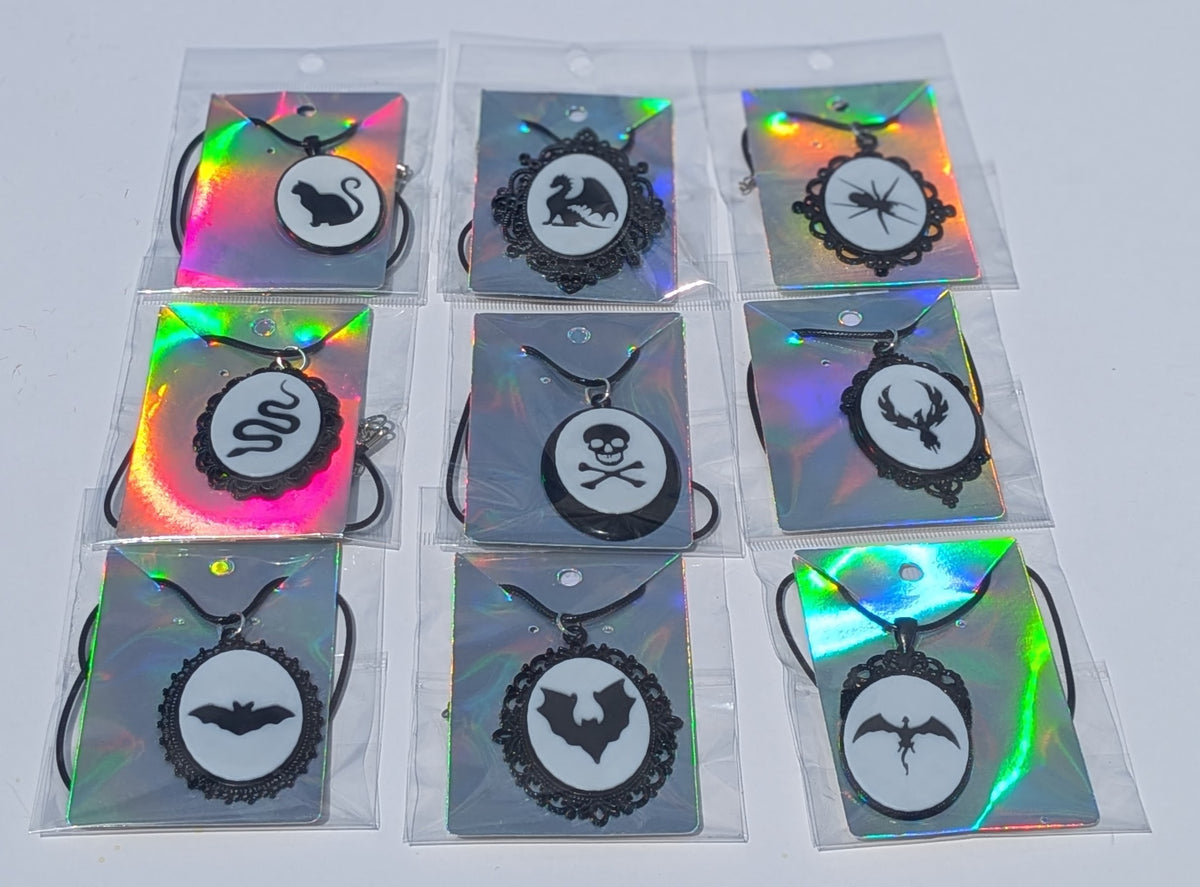 Nine gothic-themed pendant necklaces in clear plastic packaging, featuring black silhouettes of a swan, a snake, a spider, a skull and crossbones, a stag, a bat, and a winged emblem on holographic backing cards.