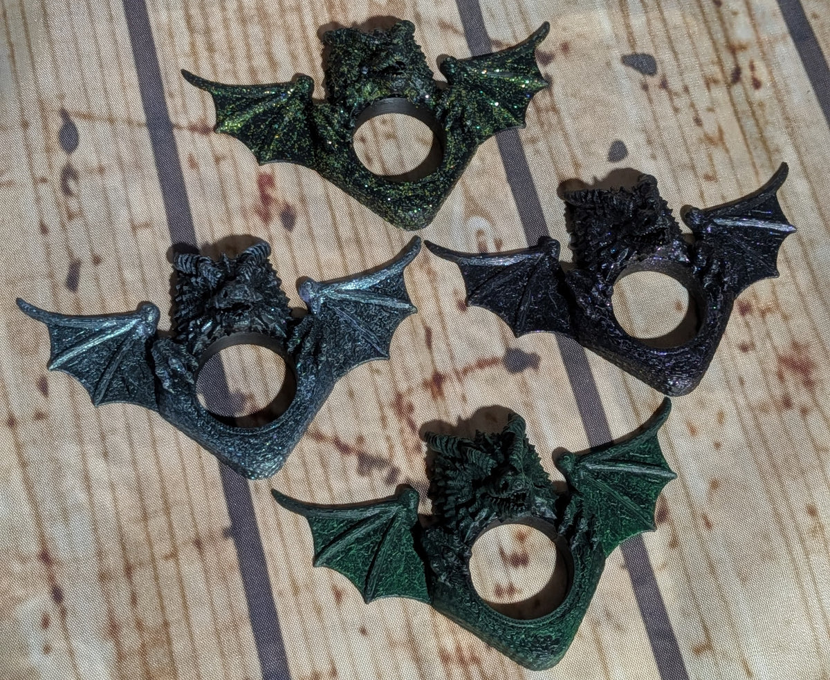 Four bat-shaped ring holders with wings spread, shown in dark metallic and textured finishes on a wood-grain background.