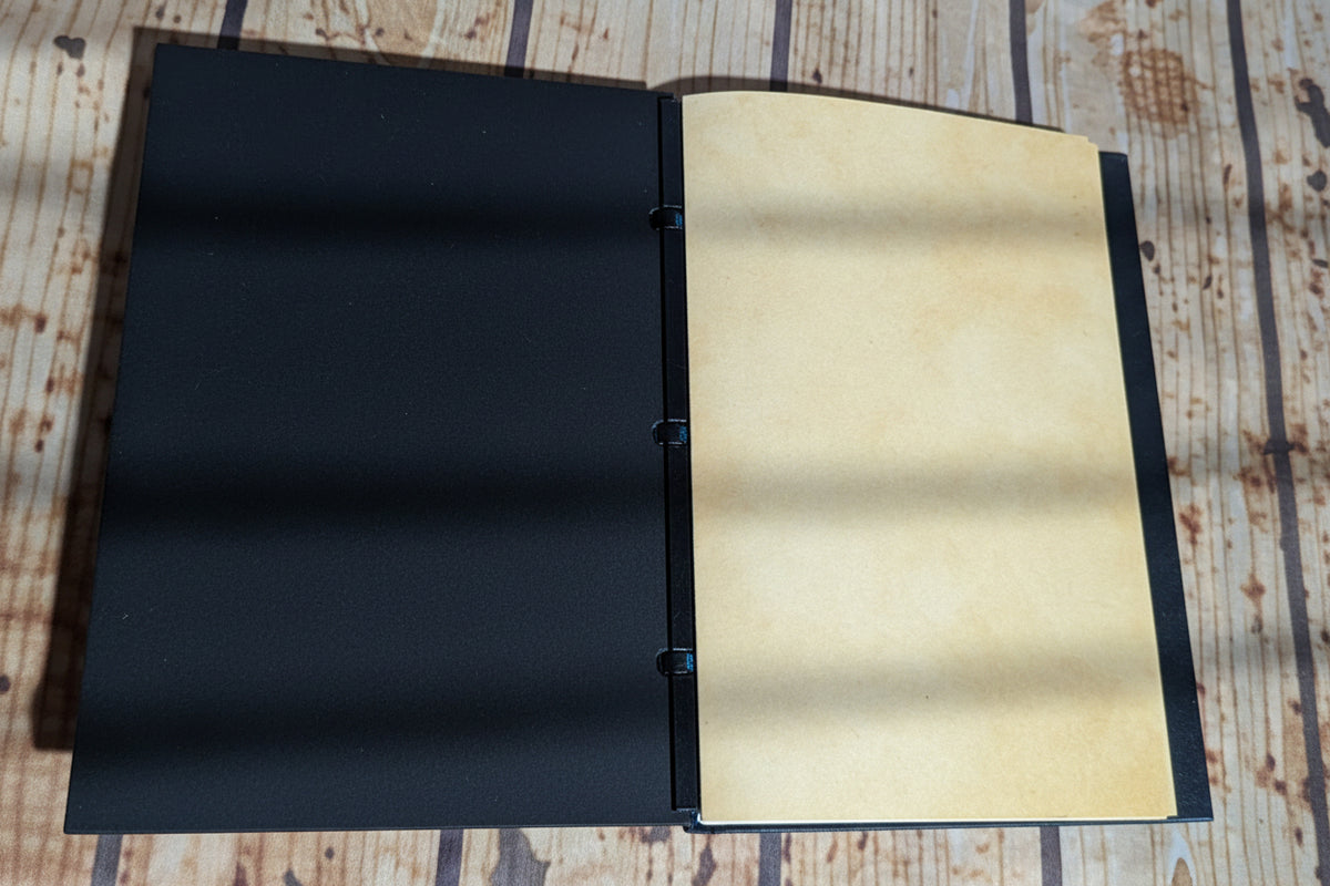 An open handmade ring-bundled journal with a black cloth cover and blank beige paper pages, displayed on a rustic wooden surface.