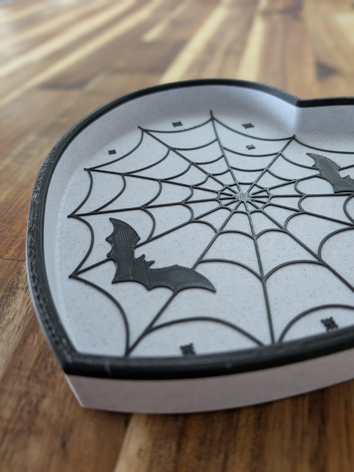 A heart-shaped Halloween-themed tray with a black spiderweb design and bat silhouettes, shown on a wooden tabletop.