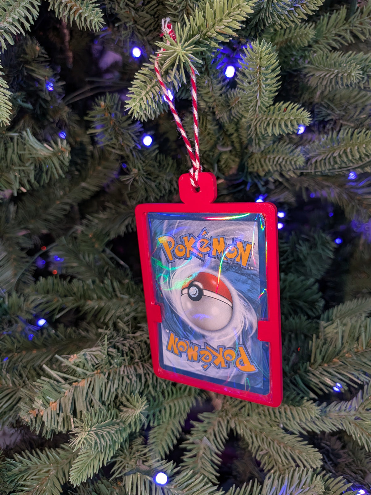A Pokémon-themed Christmas ornament shaped like a trading card with a red border and a red-and-white hanging string, hanging on a green Christmas tree with blue lights.