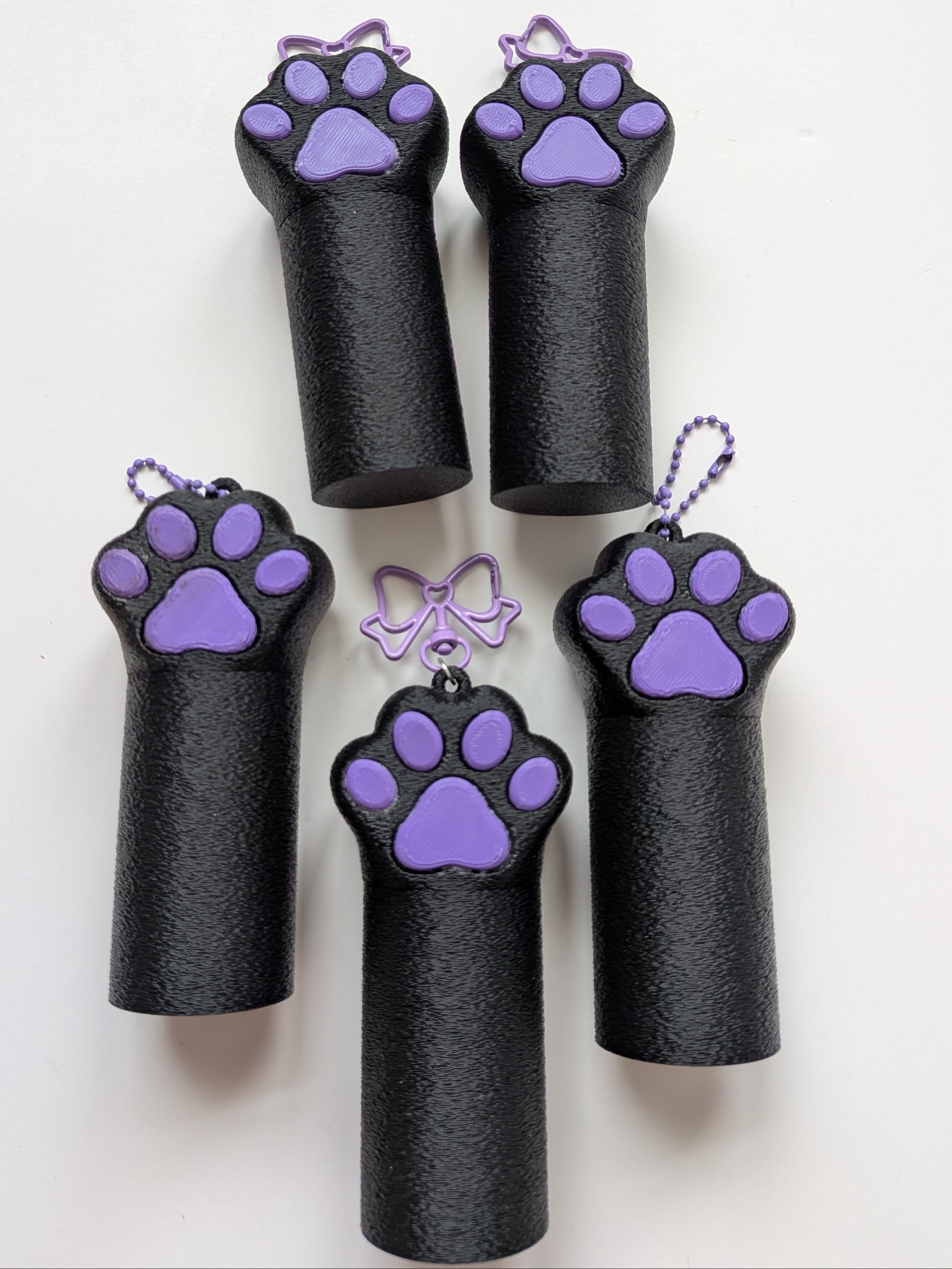Five black cylindrical chapstick bagcharms with purple paw print screw on lid hanging from purple ball chain or purple bow lobster claw keychain, arranged on a plain white background.