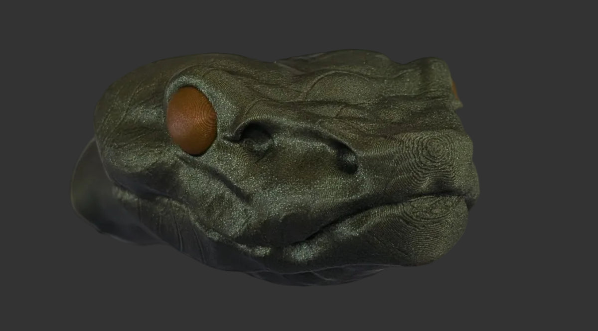 A realistic snake head figurine with orange eyes, attached to a dark gray body, shown against a printed backdrop featuring a black wire grid and a gray stone-textured background.