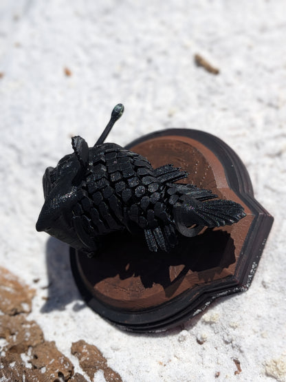 A black textured, scale-like decorative object resting on a brown and black heart-shaped base, photographed outdoors on a light sandy surface.