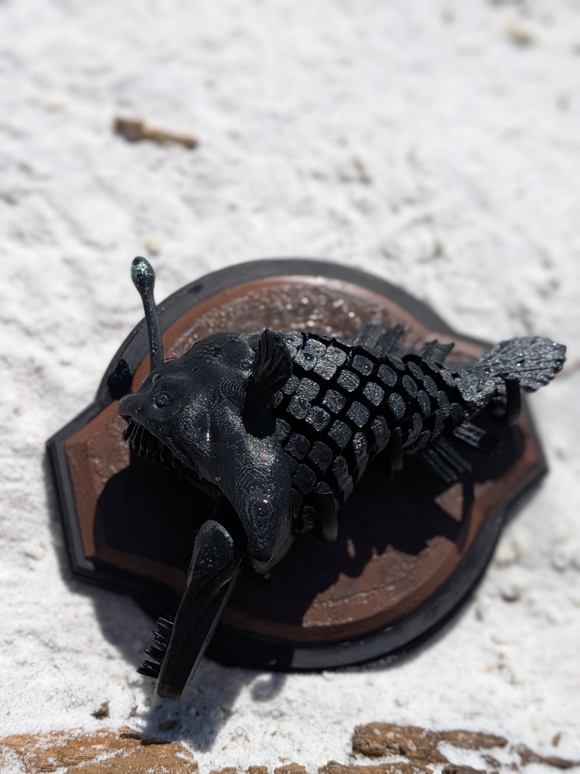 A black metal fish wall plaque featuring a detailed deep-sea anglerfish mounted on a brown shield-shaped backing, set against a white textured stone background.