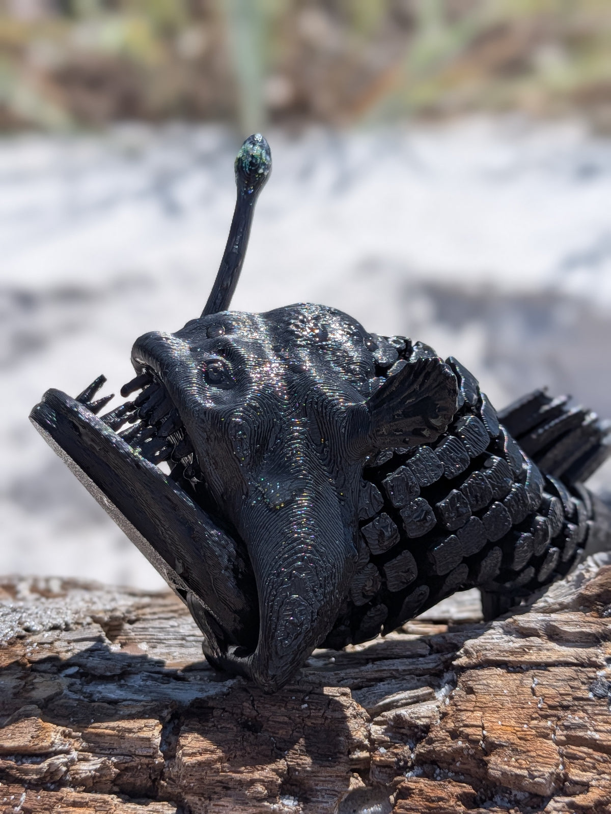 A black anglerfish sculpture with a textured, glossy finish, resting on a piece of weathered wood with a blurred natural background.