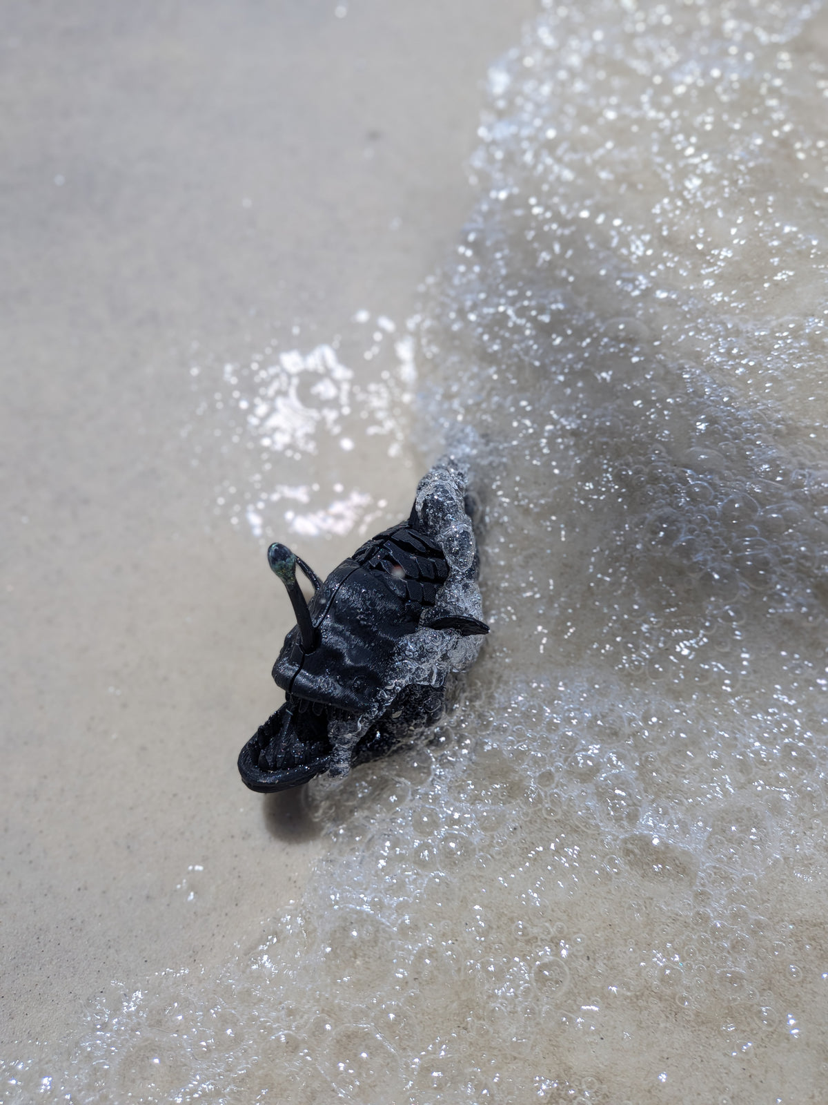 A black leather shoe or boot partially submerged in shallow ocean water with foamy surf on a sandy beach.