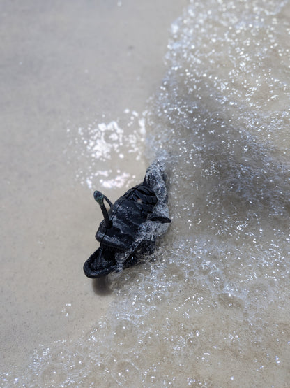 A black leather shoe or boot partially submerged in shallow ocean water with foamy surf on a sandy beach.