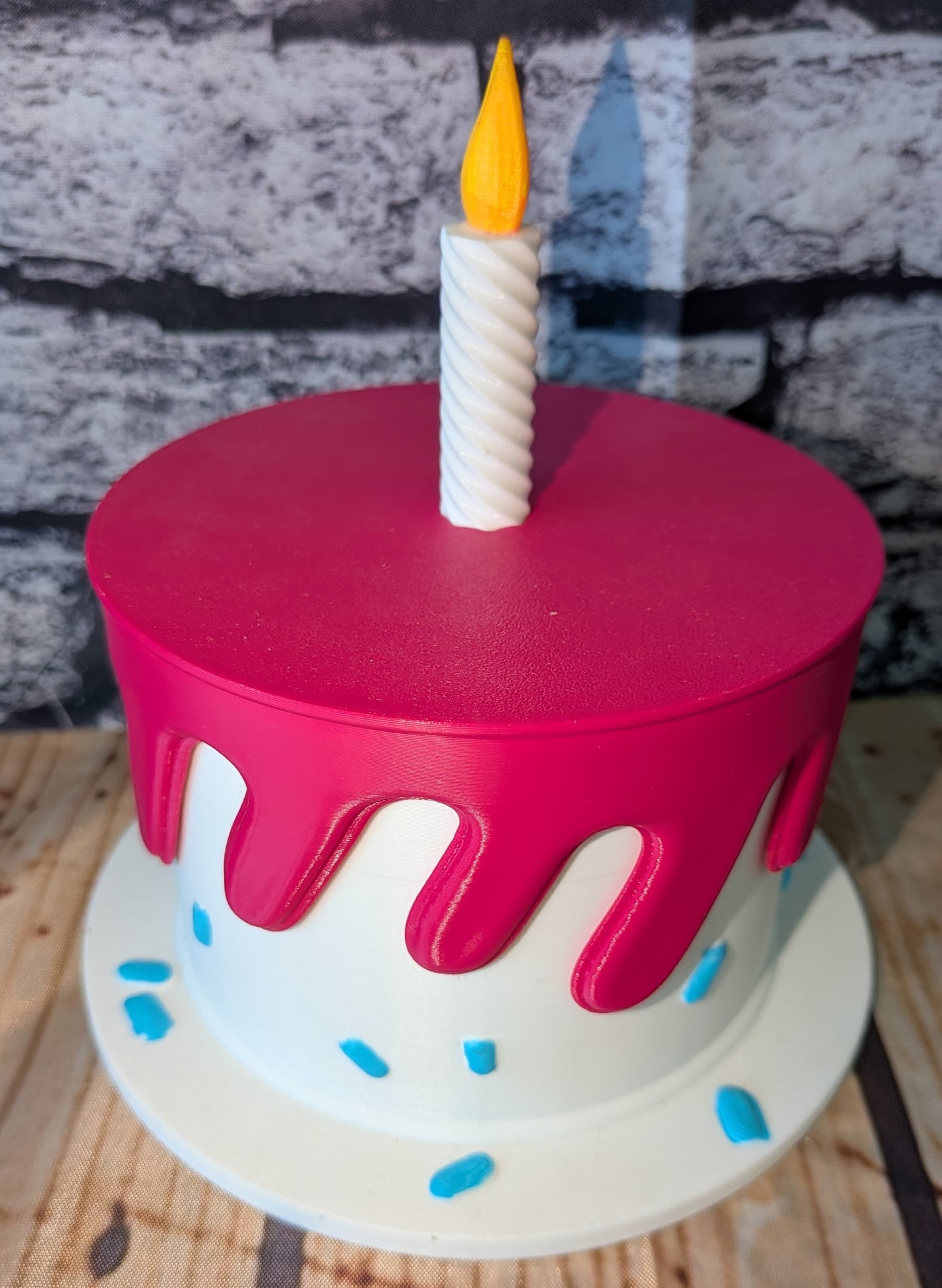 A round birthday cake with a bright pink frosting top and dripping edges, a white base, blue frosting sprinkles, and a single white spiral candle with a yellow flame on top, set against a gray brick wall background.