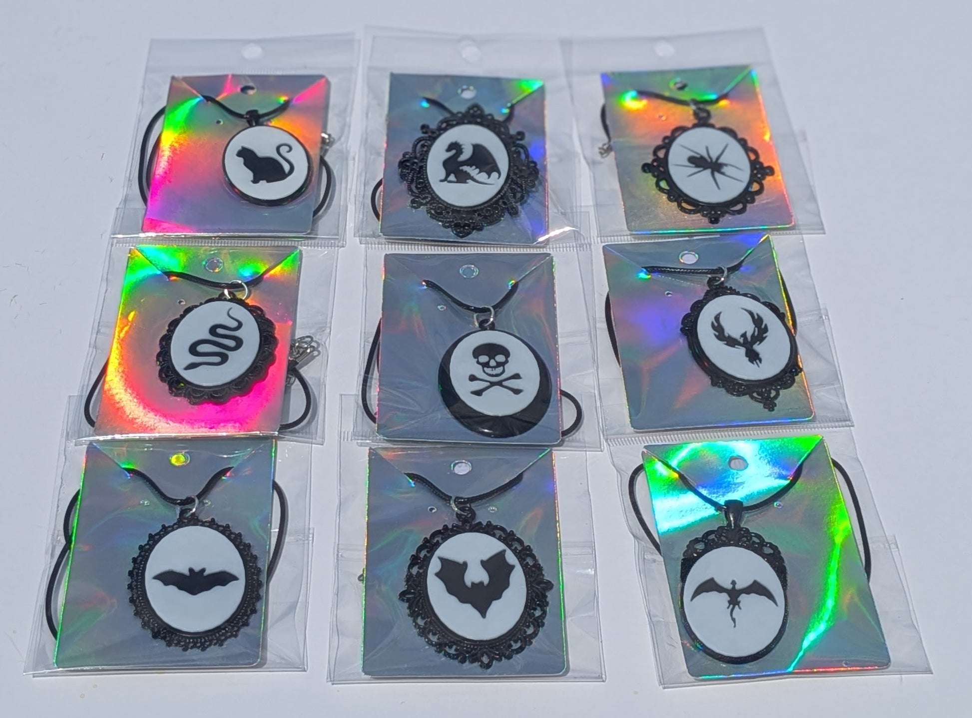 Nine gothic-themed pendant necklaces in clear plastic packaging, featuring black silhouettes of a swan, a snake, a spider, a skull and crossbones, a stag, a bat, and a winged emblem on holographic backing cards.