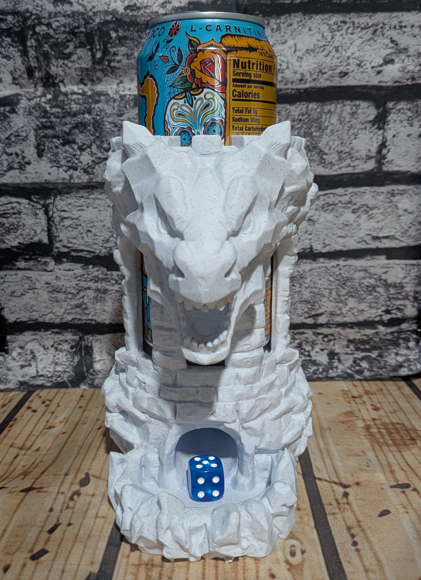 A white dragon head can holder made of stone-like material, shown holding an L-Carnival beverage can on top and a blue die in the base, set against a gray brick wall and wooden surface.