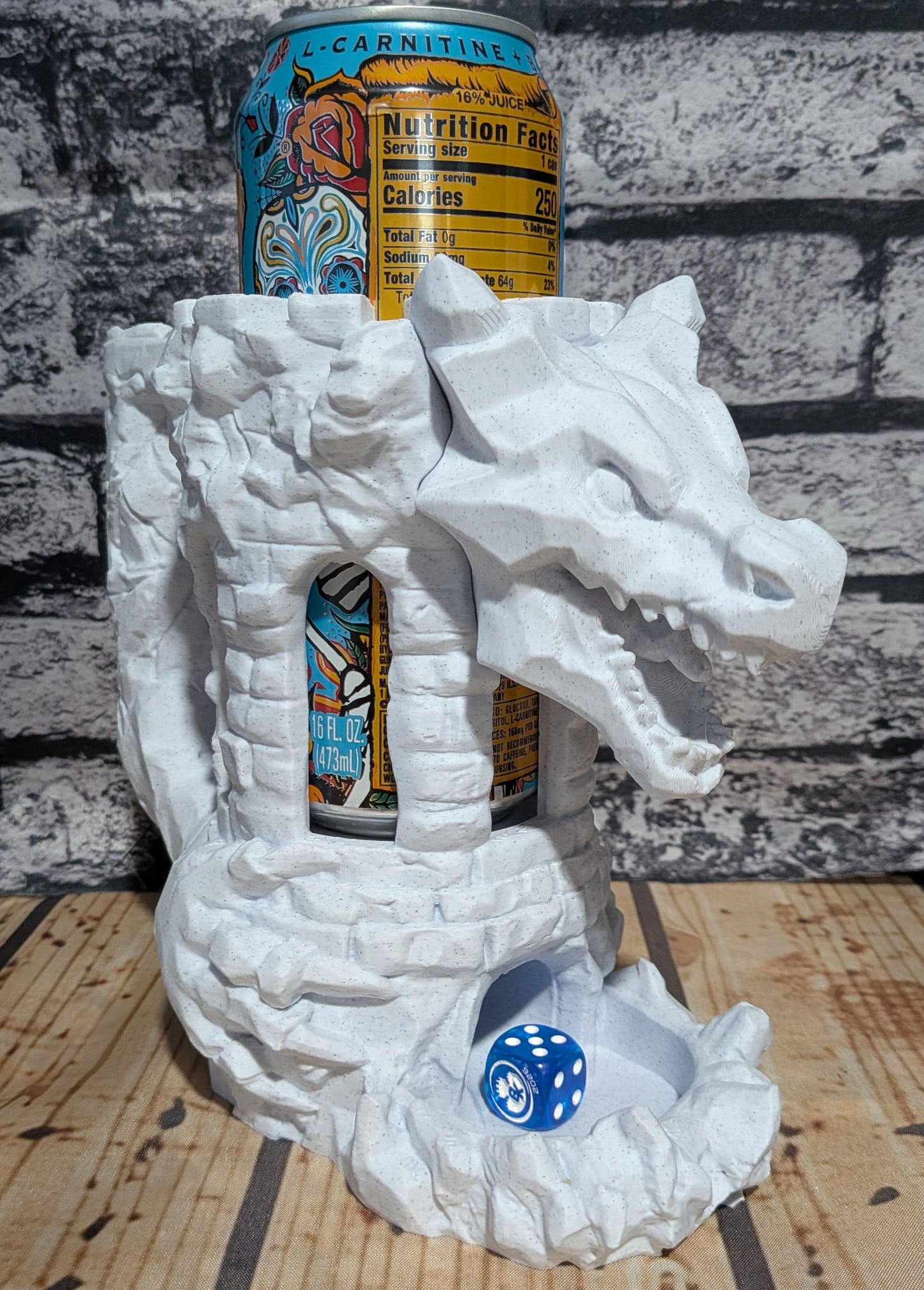 A white dragon-shaped can holder made of stone-like material, shown holding two beverage cans and a blue die on a wooden surface against a gray brick wall background.