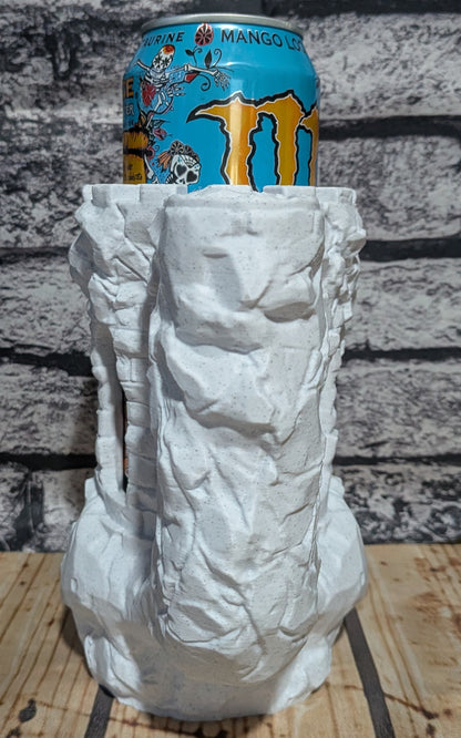 A white, highly textured sculptural can holder designed to look like a weathered stone or rock face, shown holding a blue Monster Energy drink can. The holder is placed on a wooden surface against a gray brick wall background.