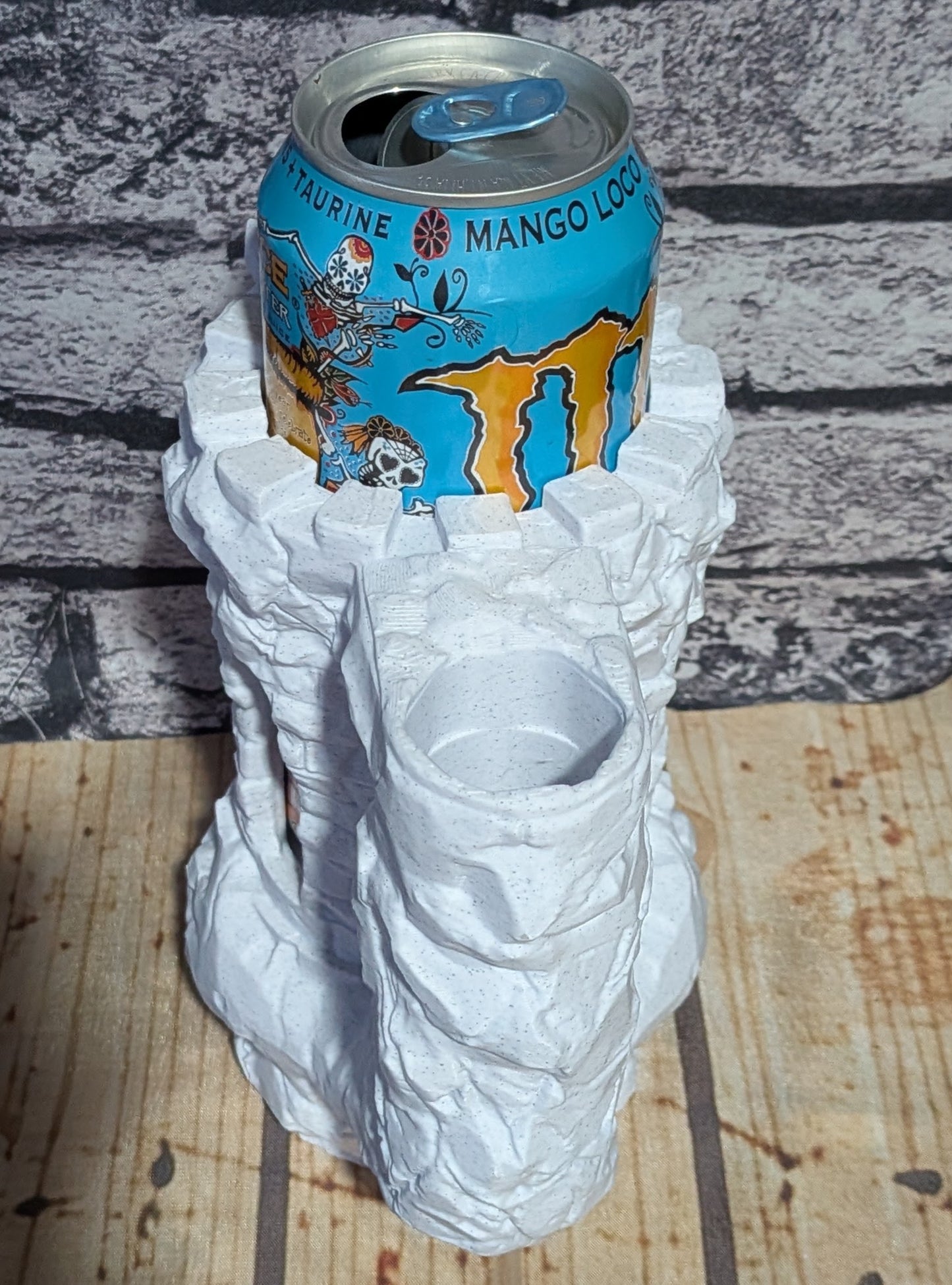 A white, crumpled-paper style can holder shaped like a rock or textured sculpture, shown holding a blue Monster Energy drink can on a wooden surface against a gray brick-patterned background.