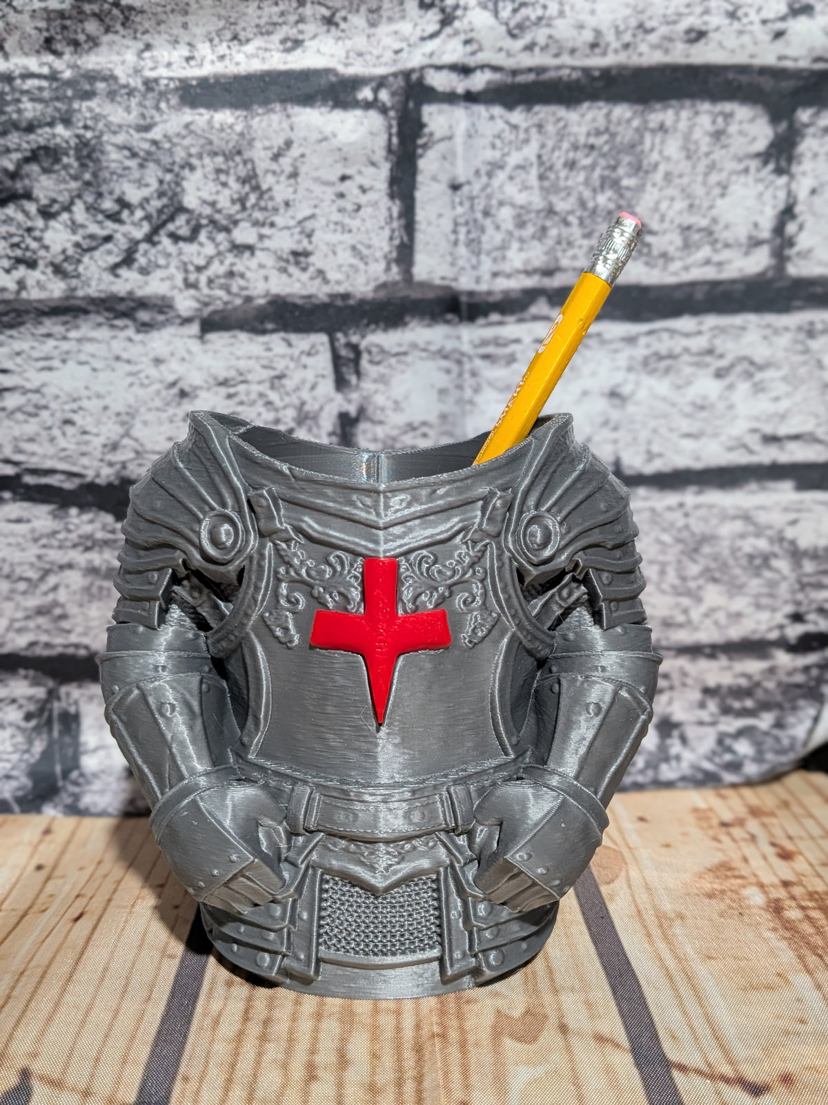 A 3D-printed knight armor desk holder in gray, featuring a red cross on the chest, shown holding a yellow pencil against a gray brick wall and wooden tabletop background.