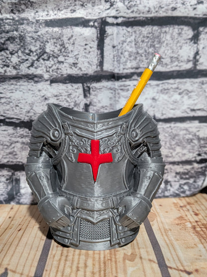 A 3D-printed knight armor desk holder in gray, featuring a red cross on the chest, shown holding a yellow pencil against a gray brick wall and wooden tabletop background.