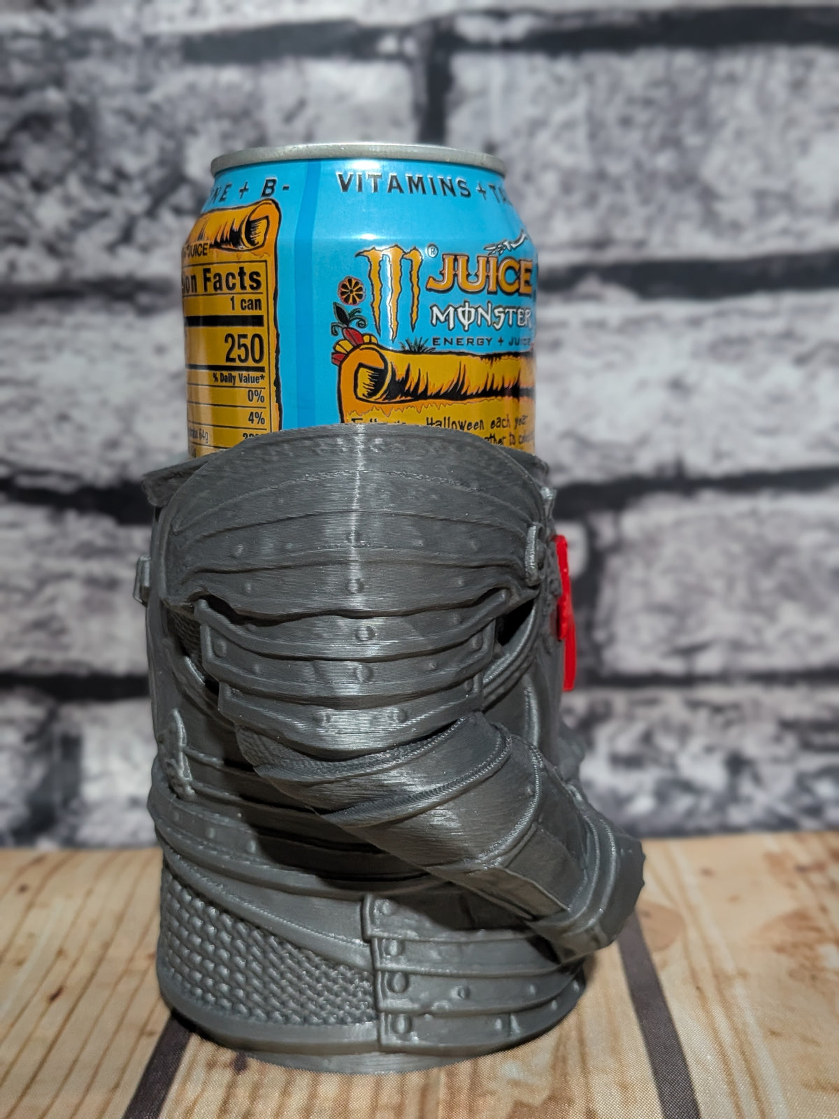 A 3D-printed miniature monster boot holder in dark gray, shown holding a Monster Energy drink can on a wooden surface against a gray brick wall background.