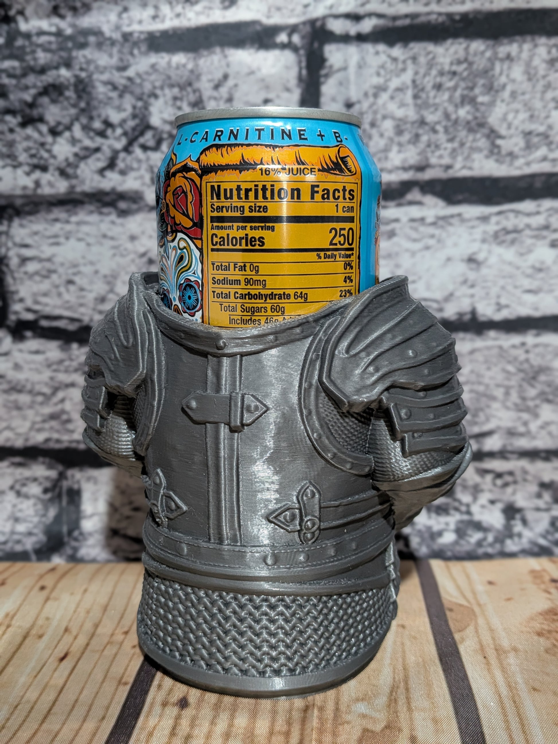 A gray 3D-printed miniature armor mug holding a can of Carnitine + B energy drink, set against a gray brick wall and light wood tabletop.