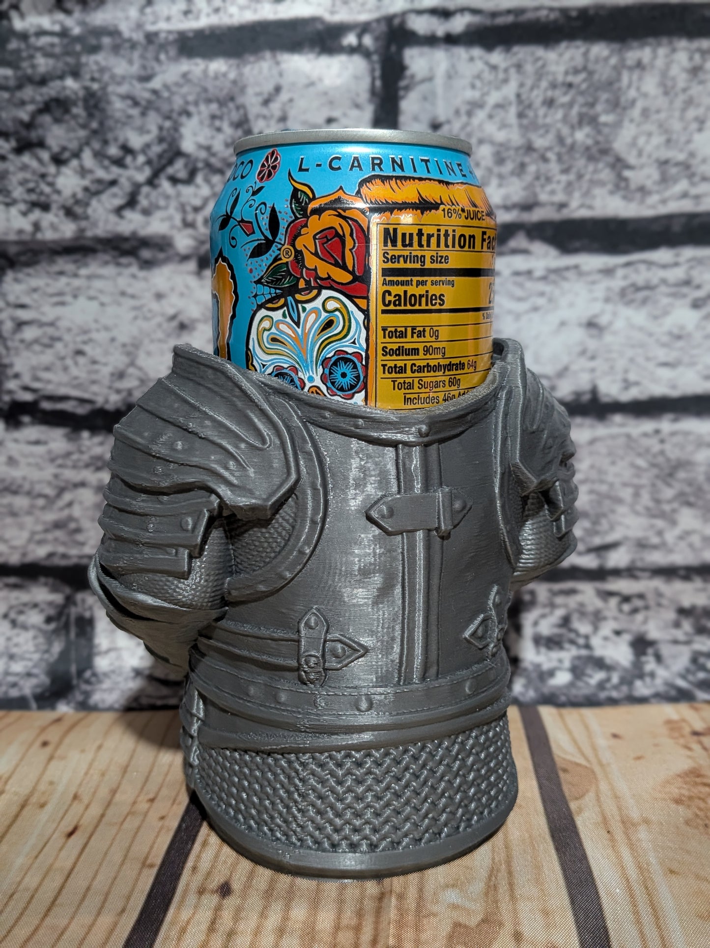 A gray 3D-printed miniature armor can cooler holding a can of L-Carnitine, set on a wooden surface against a gray brick wall background.