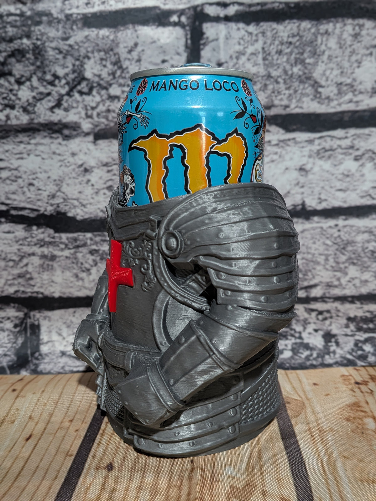 A gray 3D-printed knight helmet-shaped can holder cradling a blue Monster Energy drink can, set on a wooden surface against a gray brick wall background.