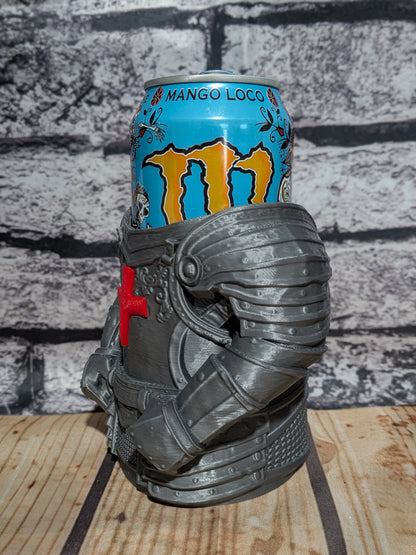 A gray 3D-printed knight helmet-shaped can holder cradling a blue Monster Energy drink can, set on a wooden surface against a gray brick wall background.
