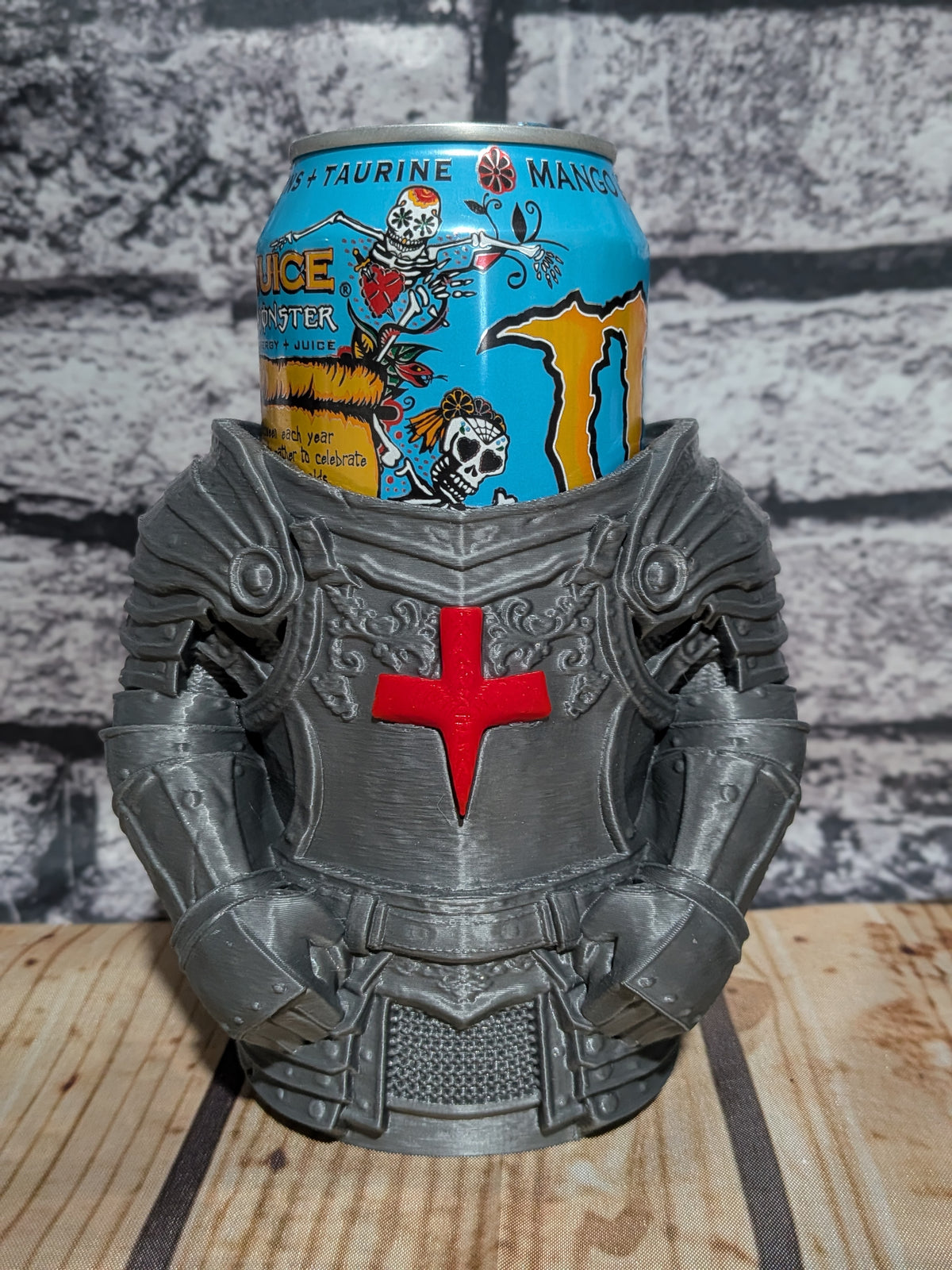 A gray knight armor mug holding a can of Monster Energy Drink, set on a wooden surface against a gray brick wall background.