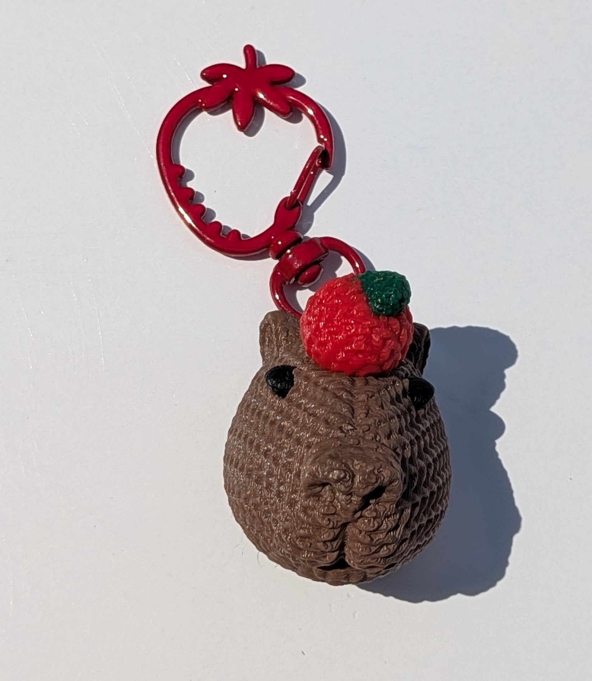 A small brown crocheted animal charm with a red strawberry on its head, attached to a red metal key ring, photographed on a plain white background.
