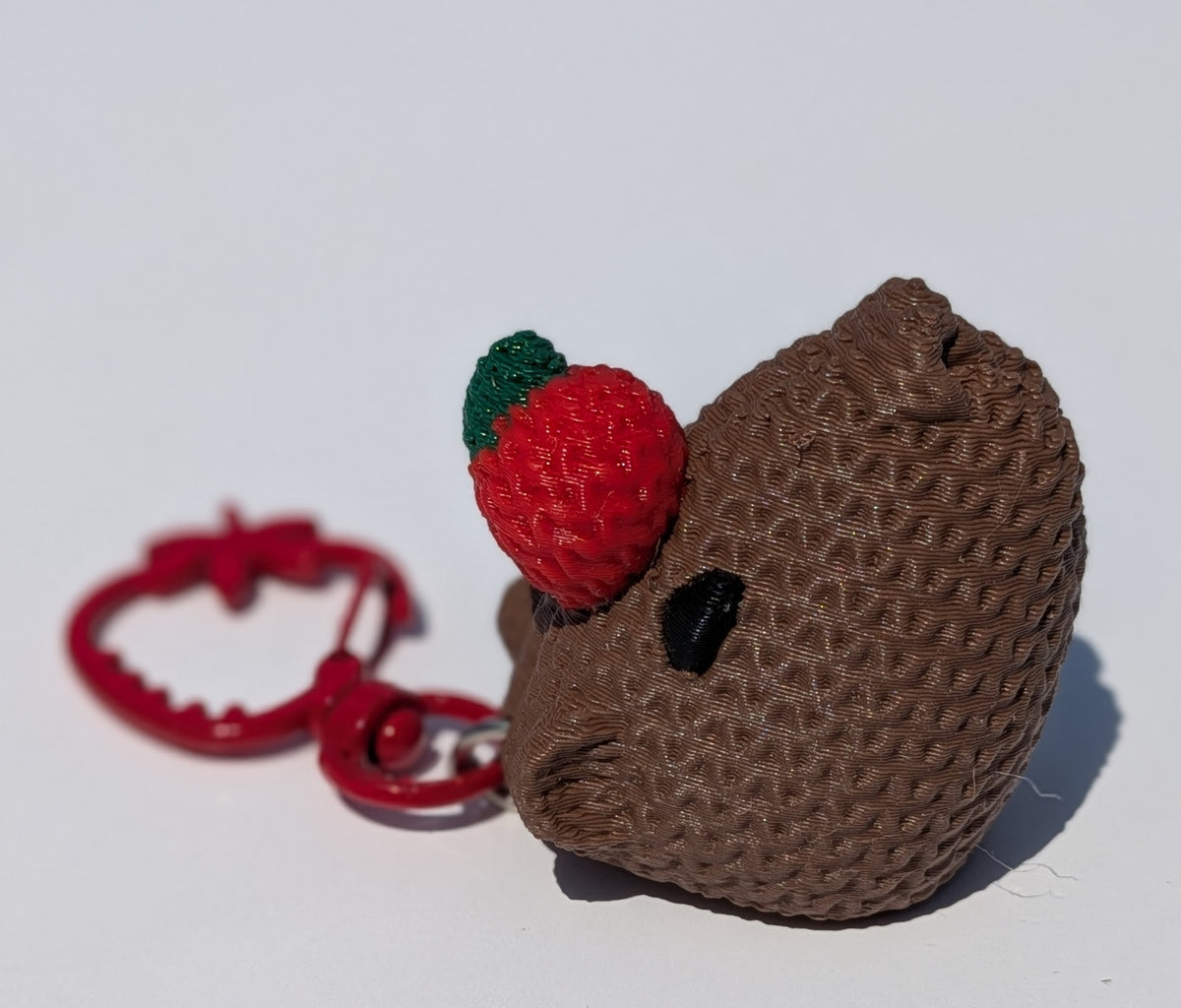 A brown textured crochet keychain charm shaped like a strawberry-topped character head, with a red heart-shaped ring and silver split ring on a plain white background.