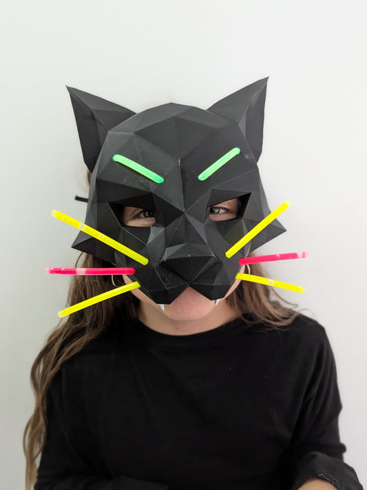 A person wearing a black low-poly cat mask with glowing green eyes and yellow, pink, and yellow whiskers, against a plain white background.