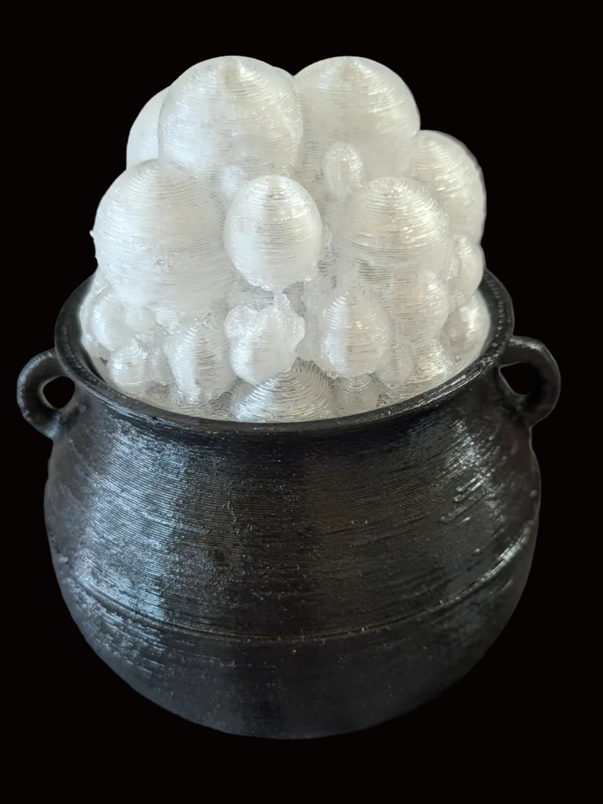 A black cauldron-shaped container holding a cluster of white, bubble-like decorative pieces, placed on a mottled gray and black surface with a dark metal rack in the background.