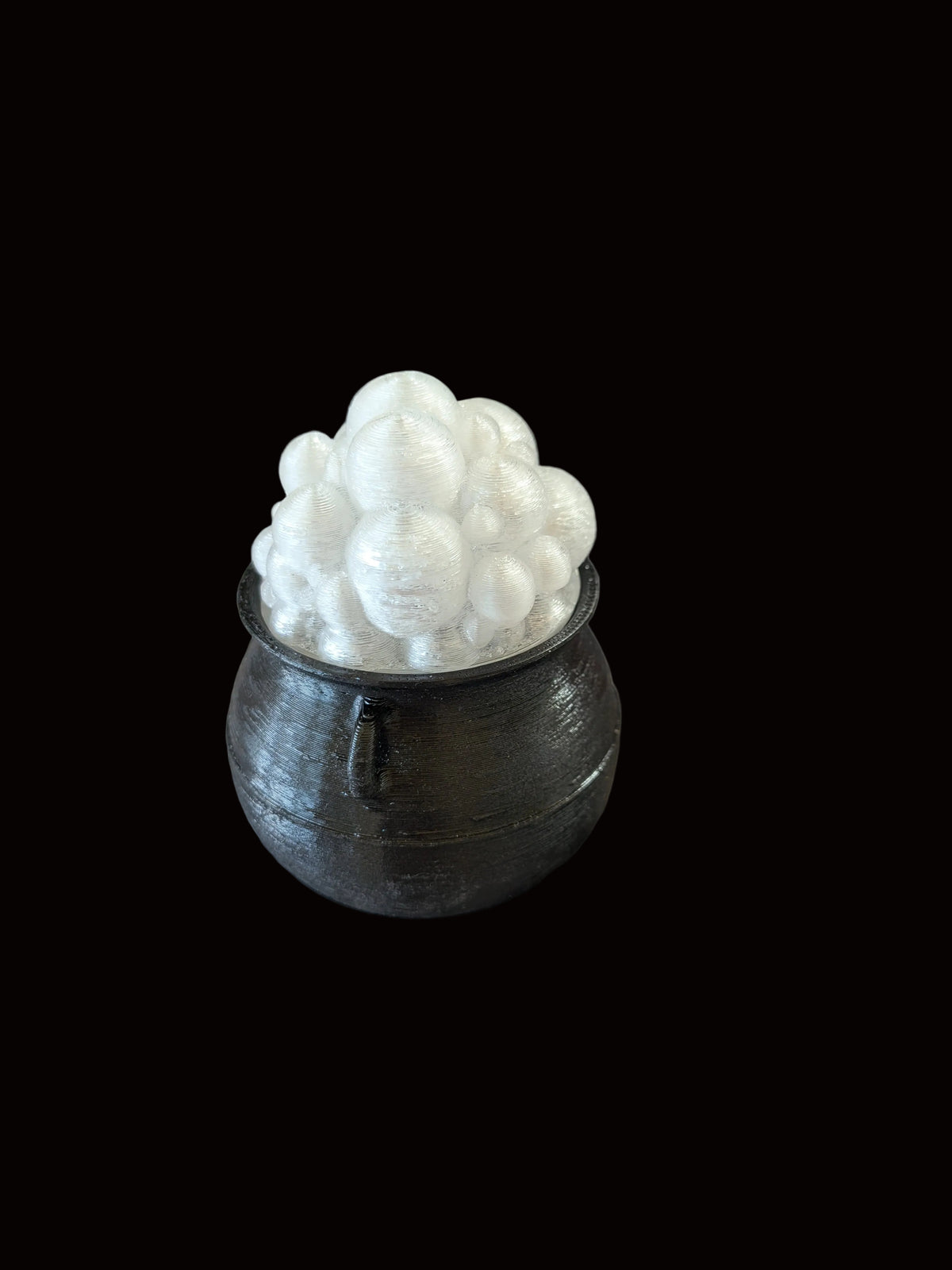 A small black cauldron-shaped container filled with white bubble-like decorative pieces, placed on a mottled gray and white surface with a dark metal rack in the background.