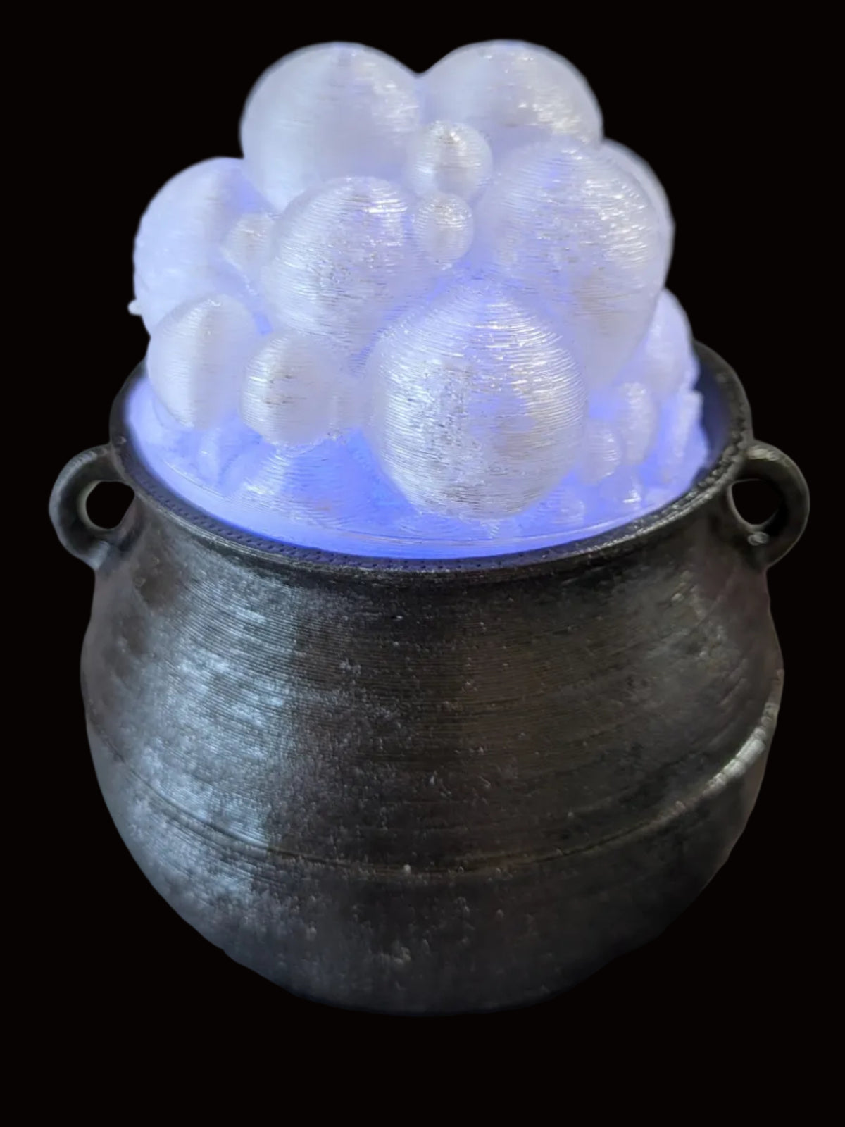 A small black cauldron-shaped container with a cluster of frosted, bubble-like white spheres inside, glowing with a blue light from within, set on a patterned surface with a dark wire rack in the background.