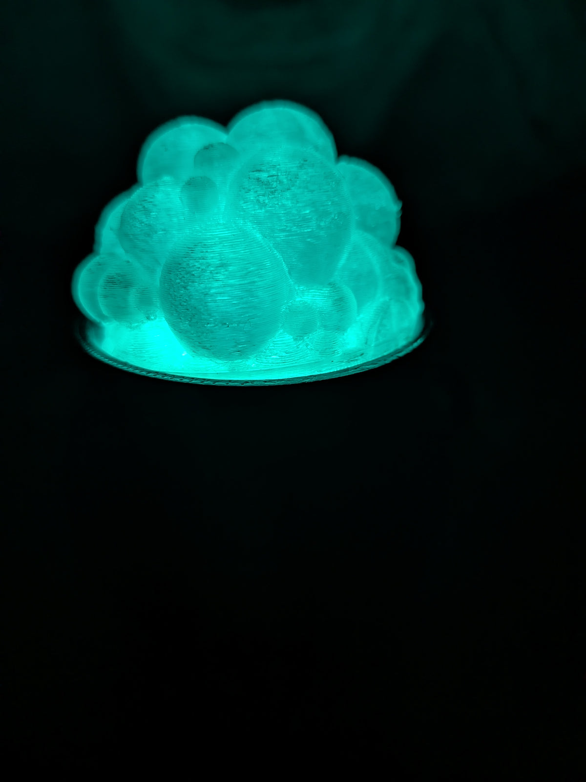 A glowing teal decorative object with a bubbled, cloud-like textured surface sitting on a dark surface against a black background.