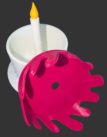 A pink plastic splatter face mask with a central hole, shown in front of a white cup containing a white twisted candle with a yellow flame, set against a dark gray background.