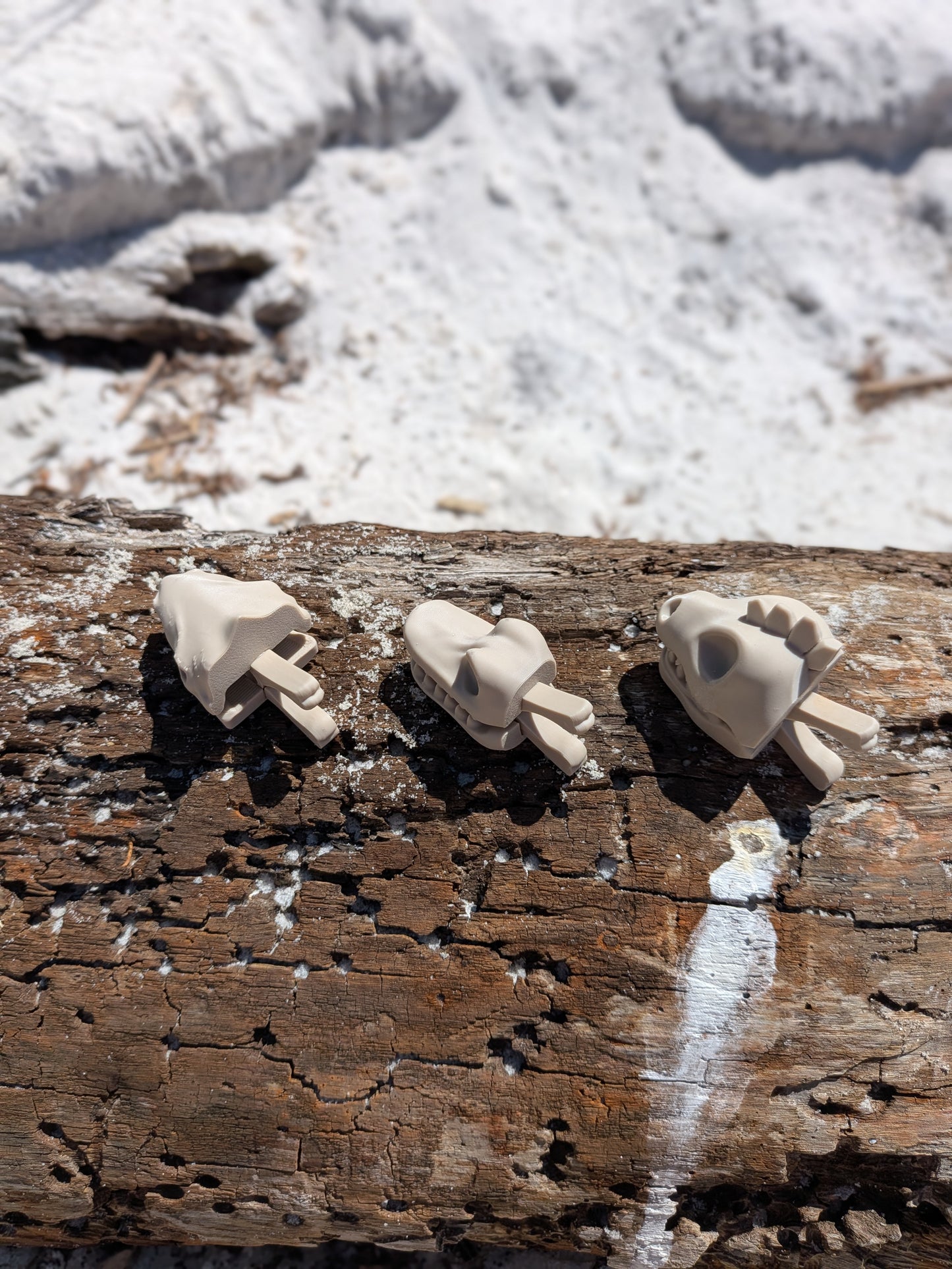 Three small beige sculptural figurines or accessories displayed on a weathered wooden log outdoors, with snow and rocks blurred in the background.
