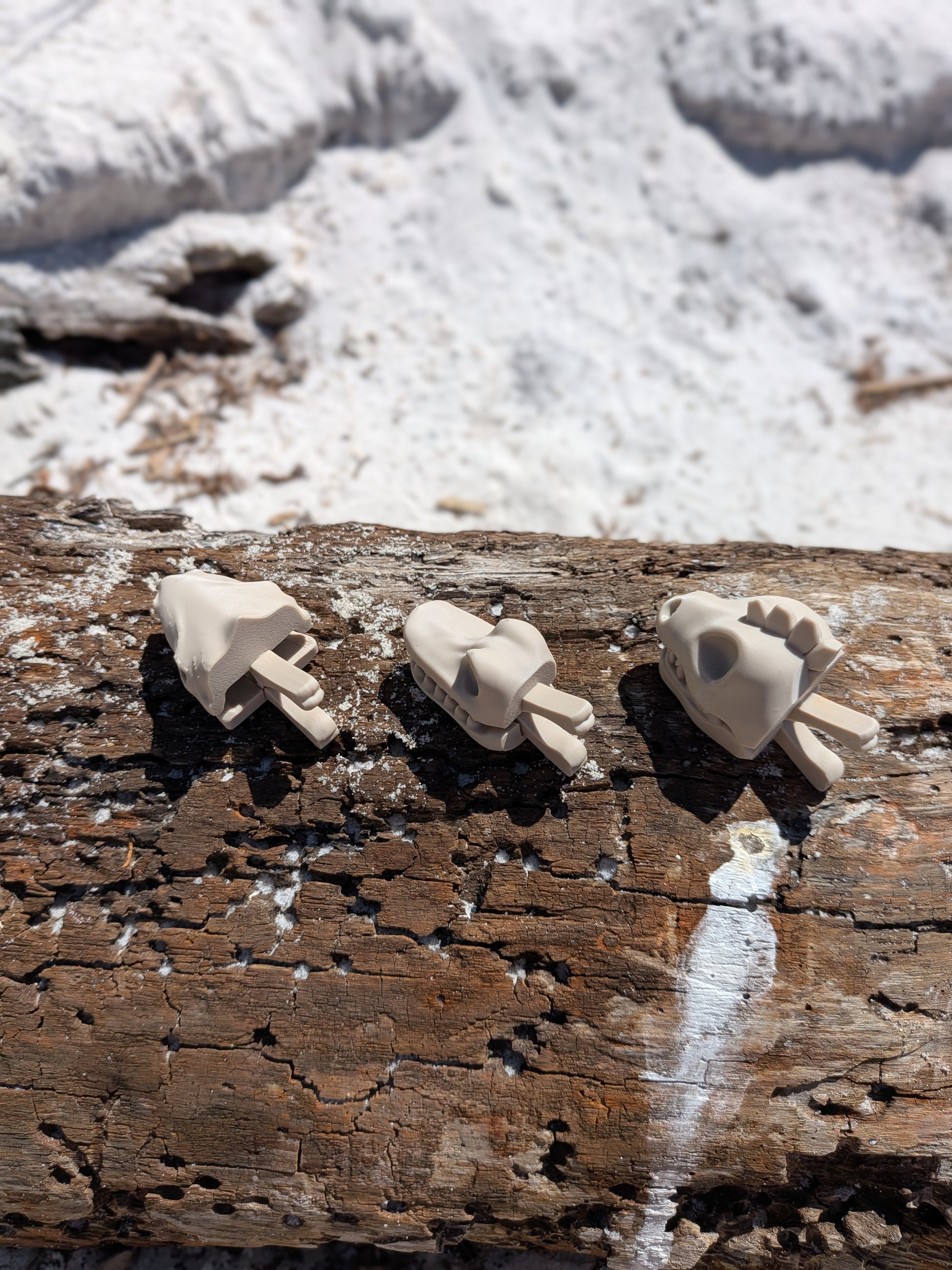 Three small beige sculptural figurines or accessories displayed on a weathered wooden log outdoors, with snow and rocks blurred in the background.