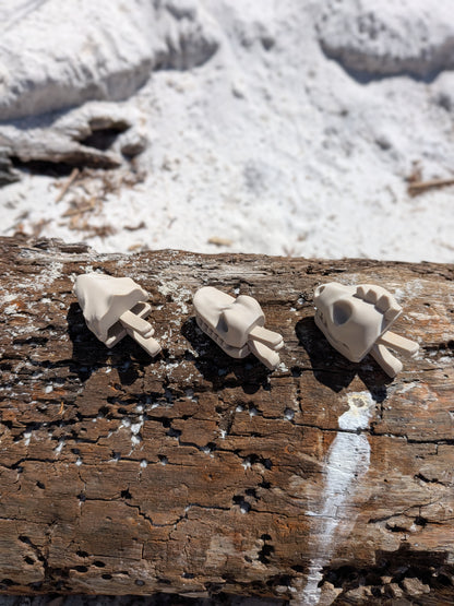 Three small beige sculptural figurines or accessories displayed on a weathered wooden log outdoors, with snow and rocks blurred in the background.