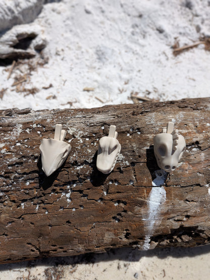 Three small beige sculptural animal skull figurines displayed on a weathered log outdoors with sand and water in the background.