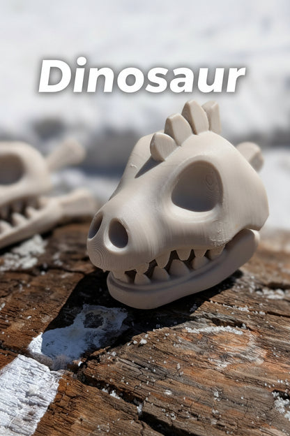 A small beige dinosaur skull figurine with sharp teeth, resting on a weathered wooden log outdoors with snow blurred in the background.