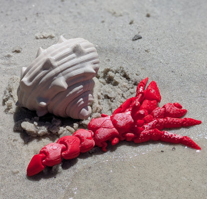 A bright red 3D-printed articulated toy crab displayed on a sandy beach next to a white seashell.