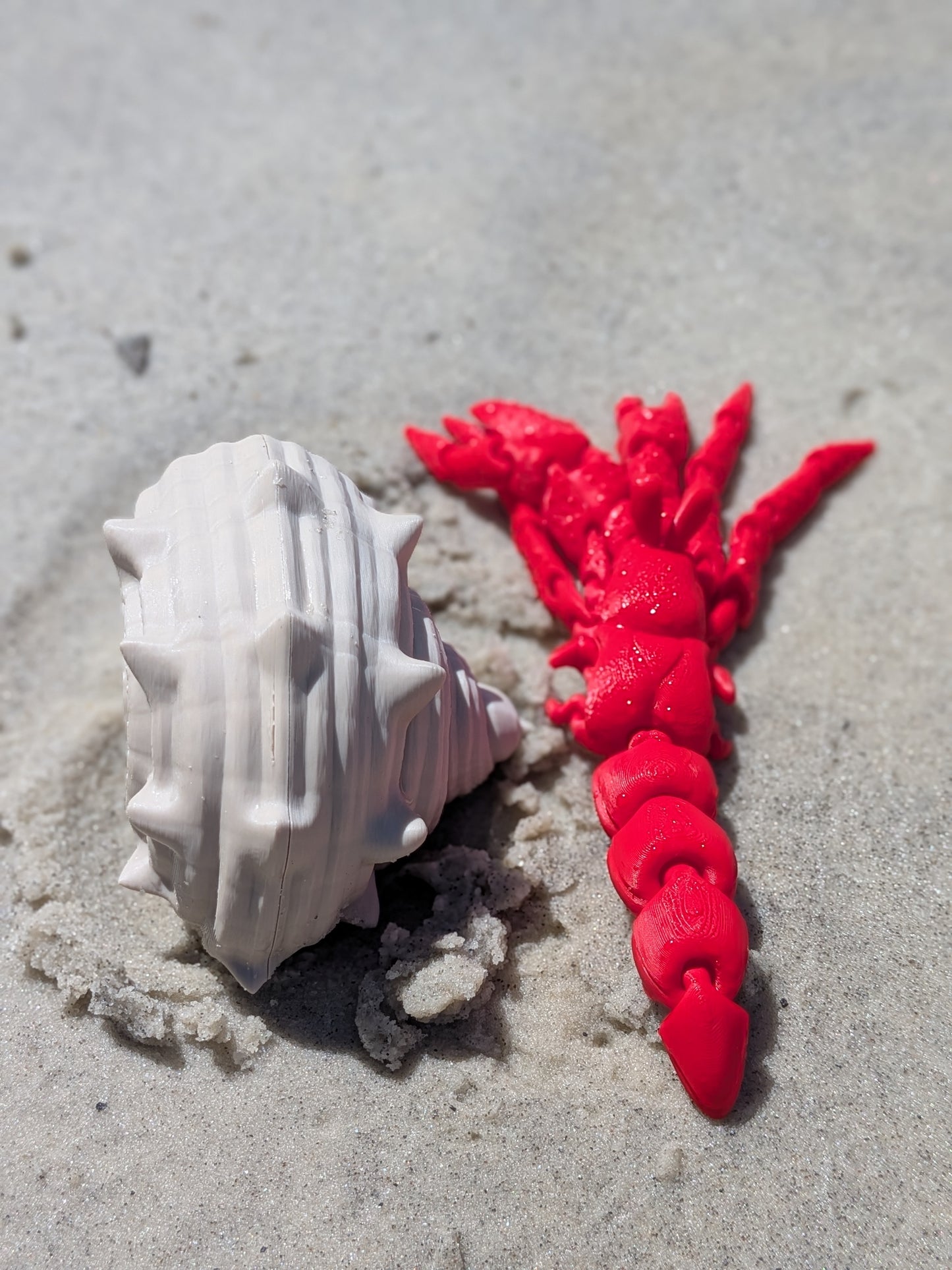 A white seashell and a bright red braided cord or hair accessory placed on light gray sand.