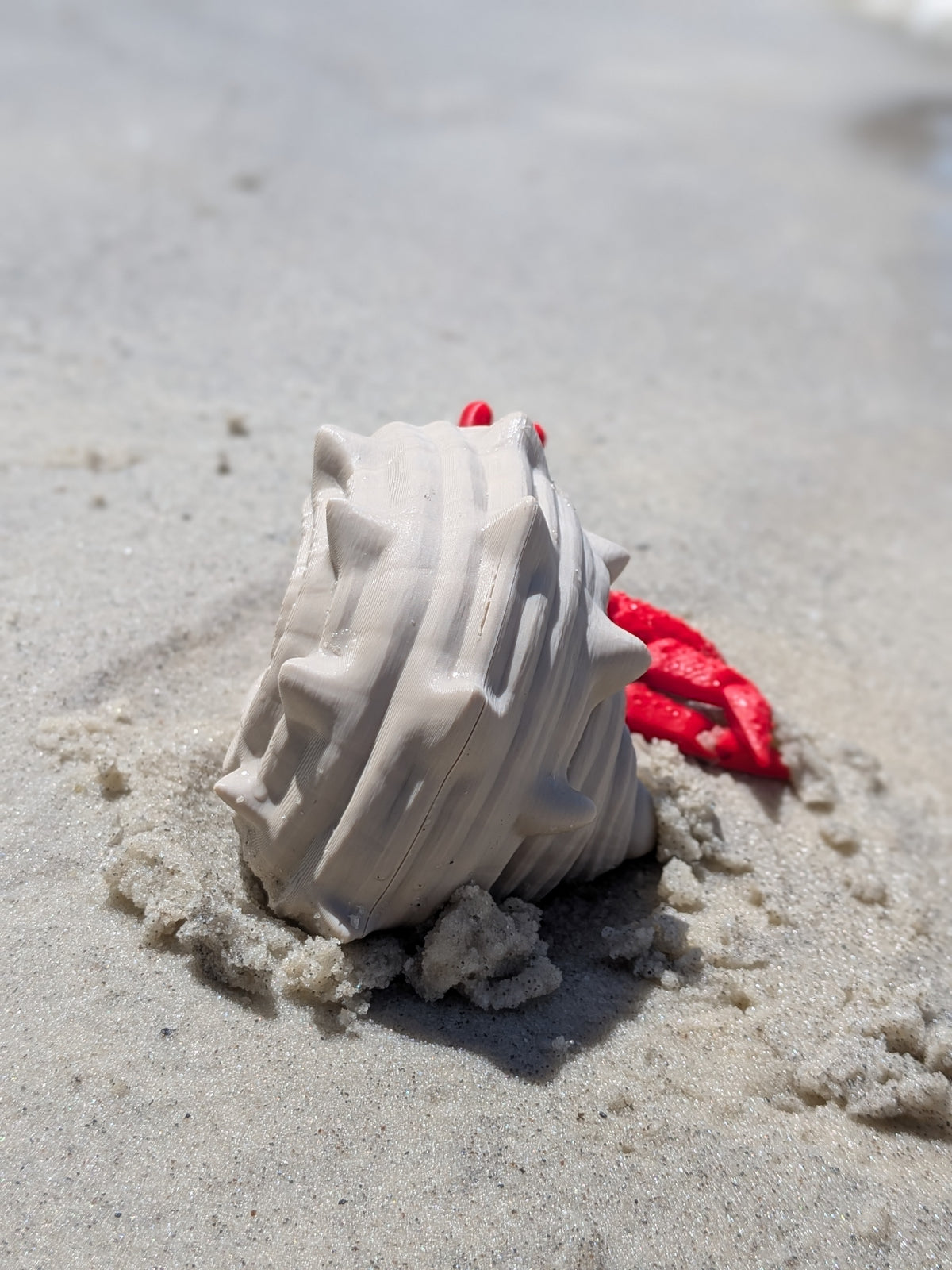 A white textured plastic seashell toy partially buried in sand on a beach, with a small red plastic shell piece visible behind it.