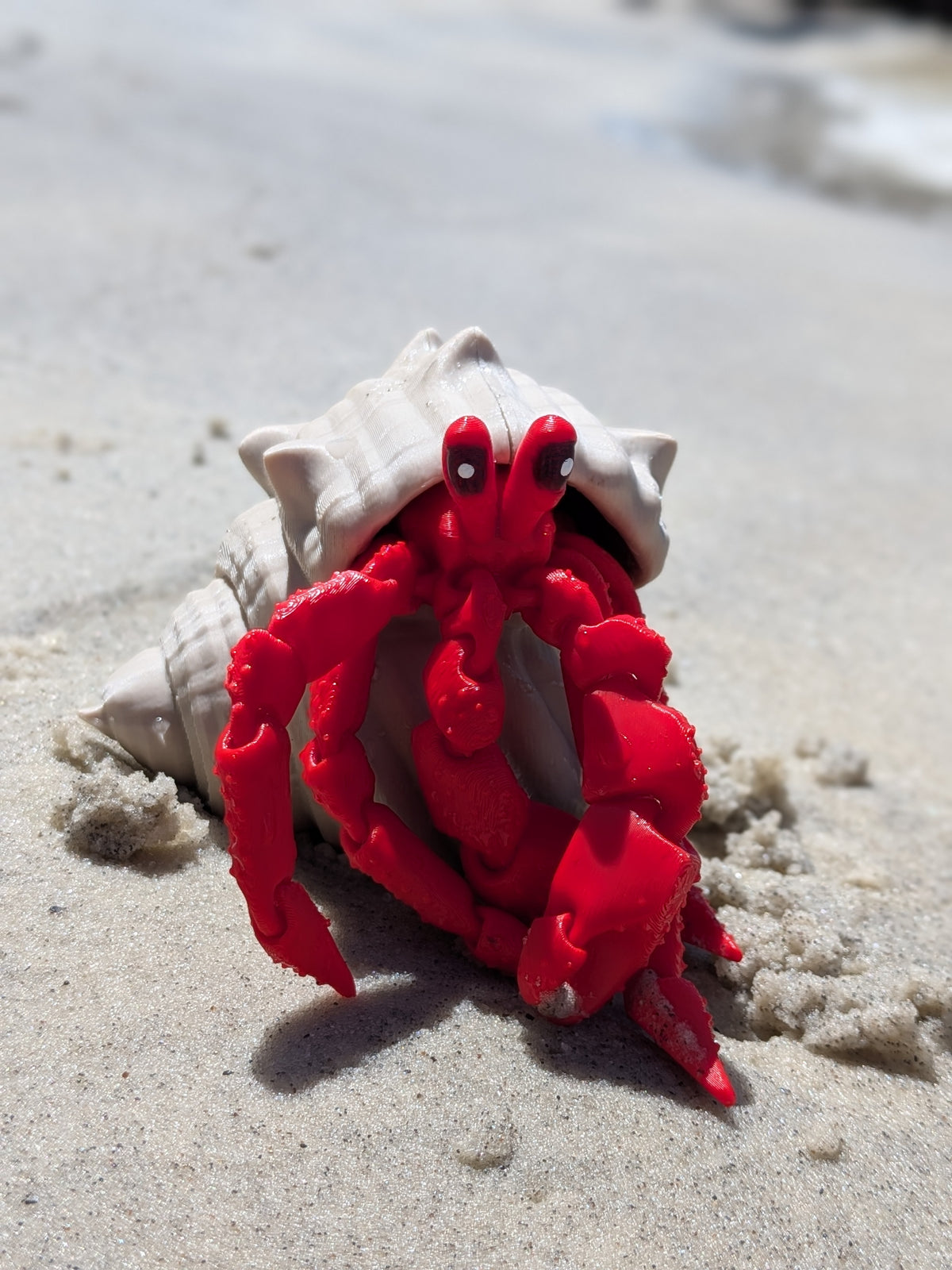 A red toy hermit crab figurine sitting in front of a white seashell on a sandy beach background.