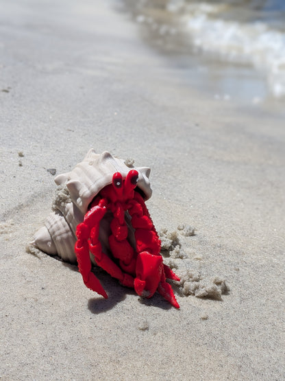 A bright red toy hermit crab figurine sitting inside a beige seashell on a sandy beach with ocean waves blurred in the background.