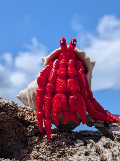 A bright red hermit crab figurine with large eyes and a beige shell, posed on a piece of driftwood outdoors against a blue sky with clouds.