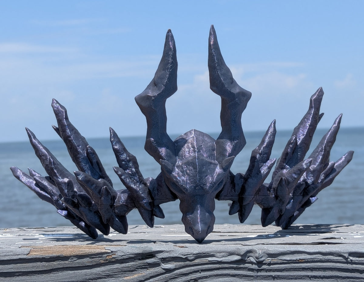 A dark metallic, horned skull sculpture or decorative head displayed outdoors on a weathered wooden surface with the ocean and blue sky in the background.
