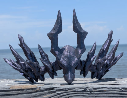 A dark metallic, horned skull sculpture or decorative head displayed outdoors on a weathered wooden surface with the ocean and blue sky in the background.