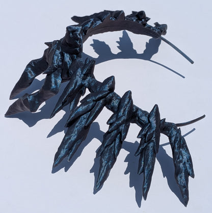 A dark metallic blue-black leaf crown headband with textured, layered leaf-like spikes, shown on a plain light background with a hard shadow.