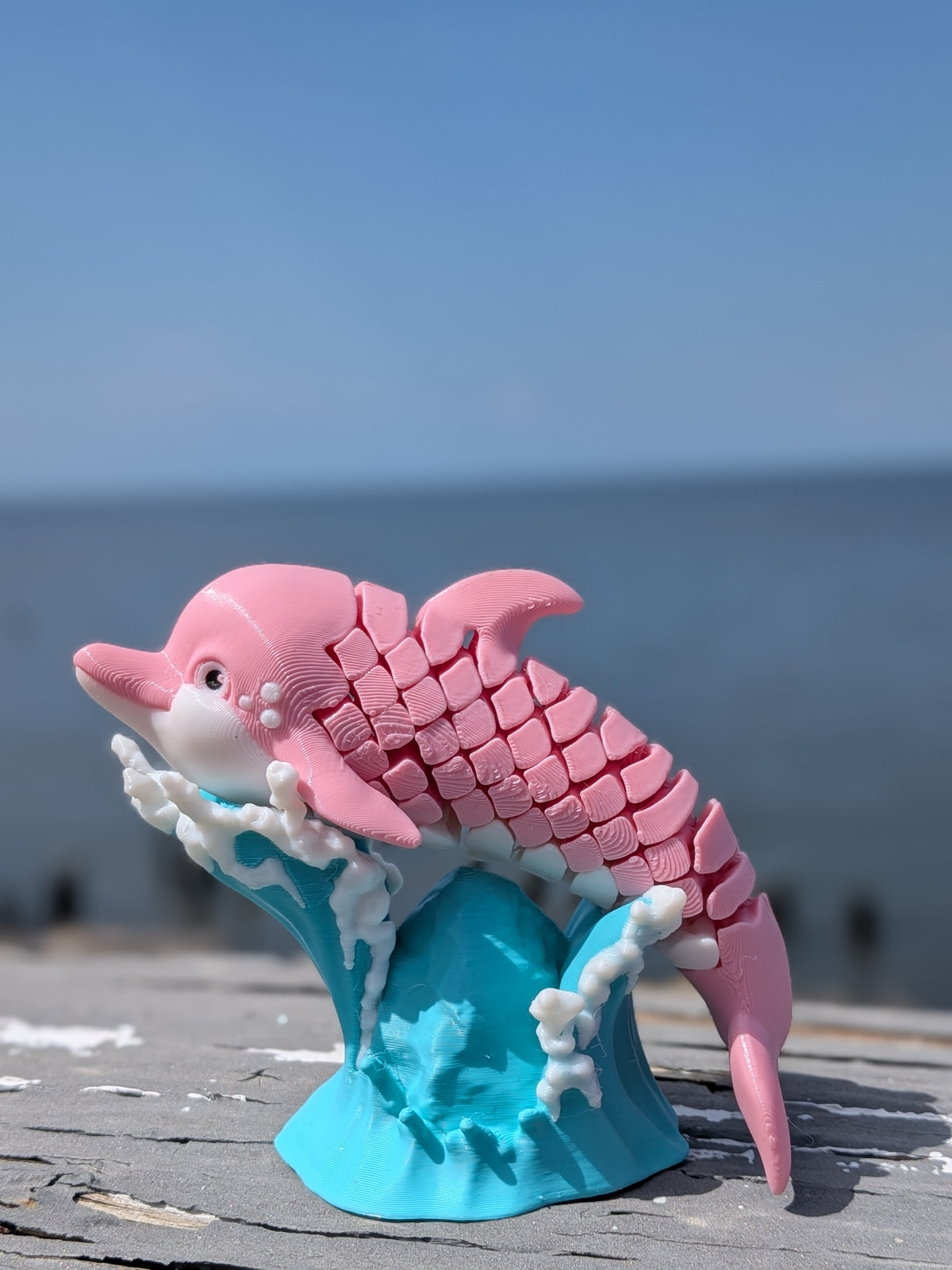 A pink 3D-printed articulated dolphin sculpture standing on a blue and white wave base, photographed outdoors on a wooden surface with the ocean and blue sky blurred in the background.