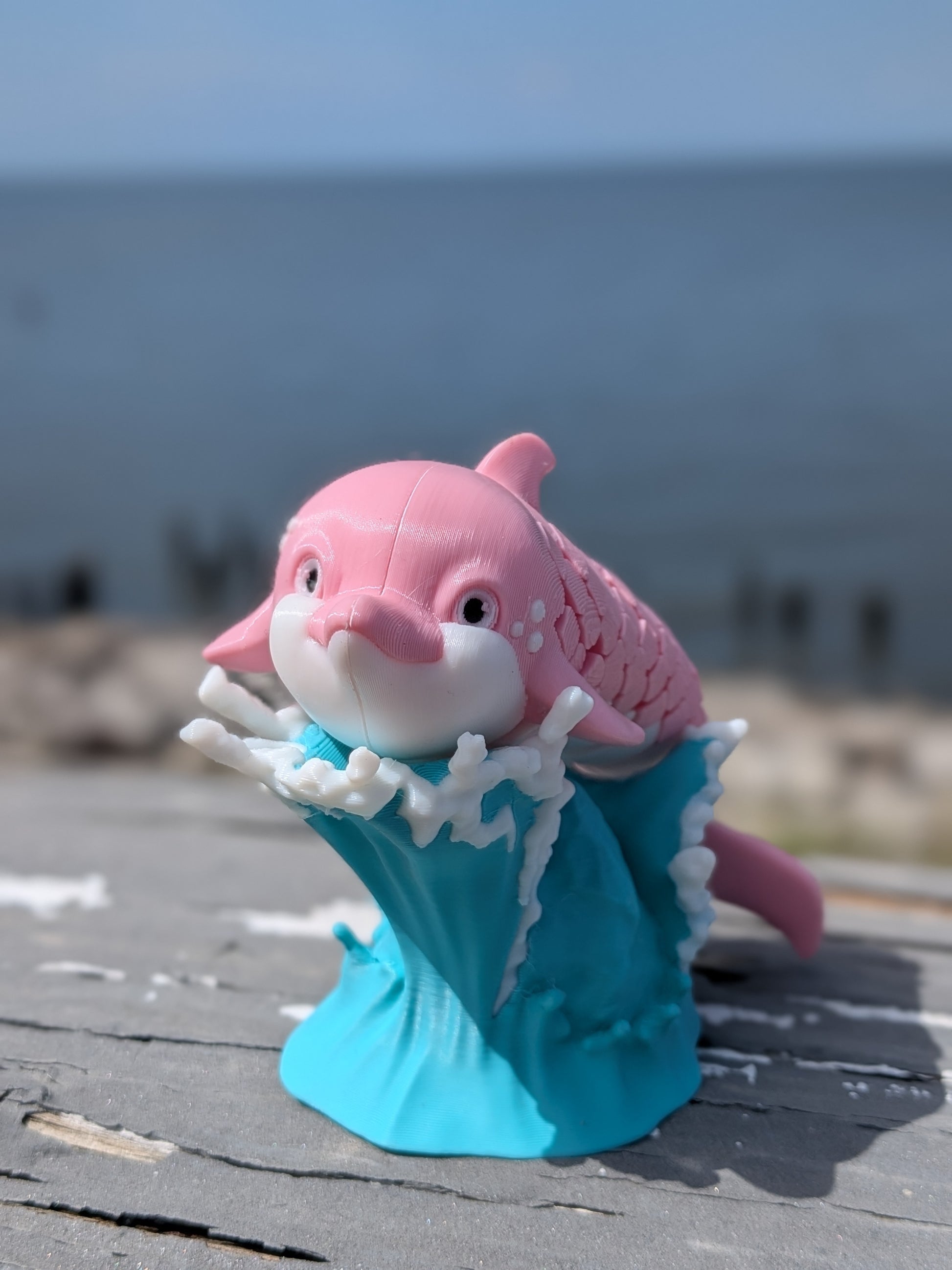 A pink and white dolphin figurine emerging from a turquoise wave base, placed on a weathered wooden surface with a blurred ocean background.